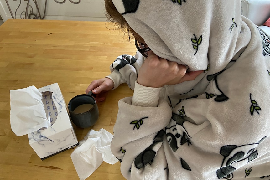 Person leaning over a table wearing a hooded gown, with a box of tissues and a mug on the table.