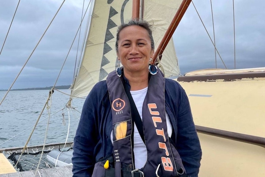 Maureen Penjueli smiling while standing on a boat with sails in the ocean. 
