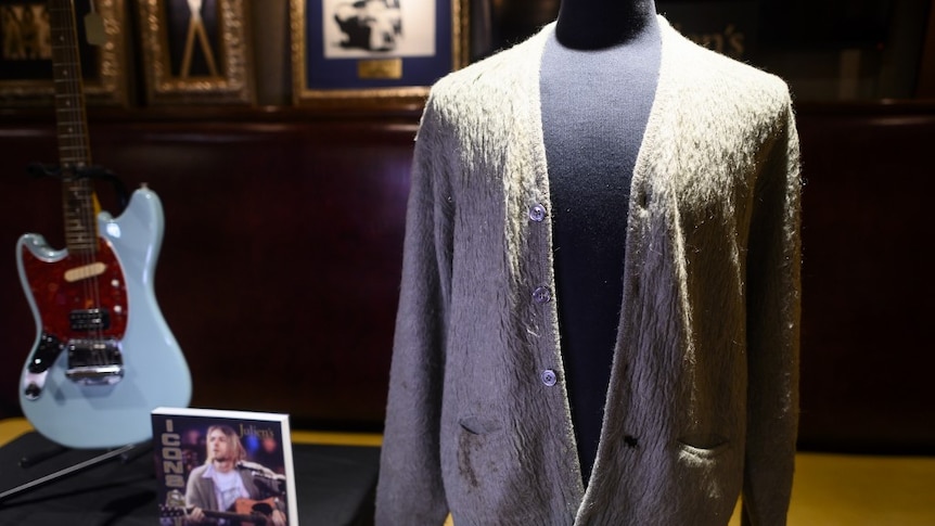 An olive green cardigan on display, in front of a book about Kurt Cobain and a guitar.