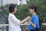 Two Japanese women with short brown hair hold hands and smile at one another on an outdoor city bridge.