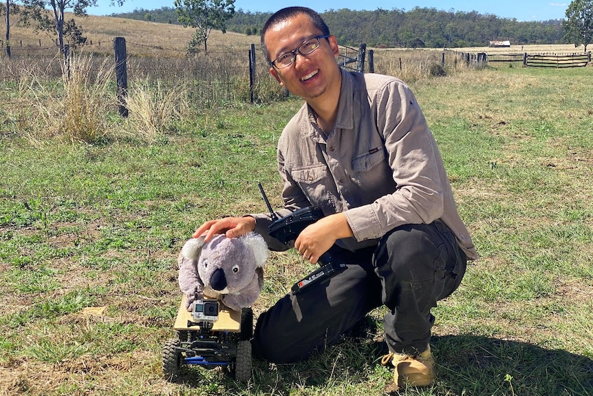 A man poses in front of a radio controlled car with a plush toy koala mounted on its top