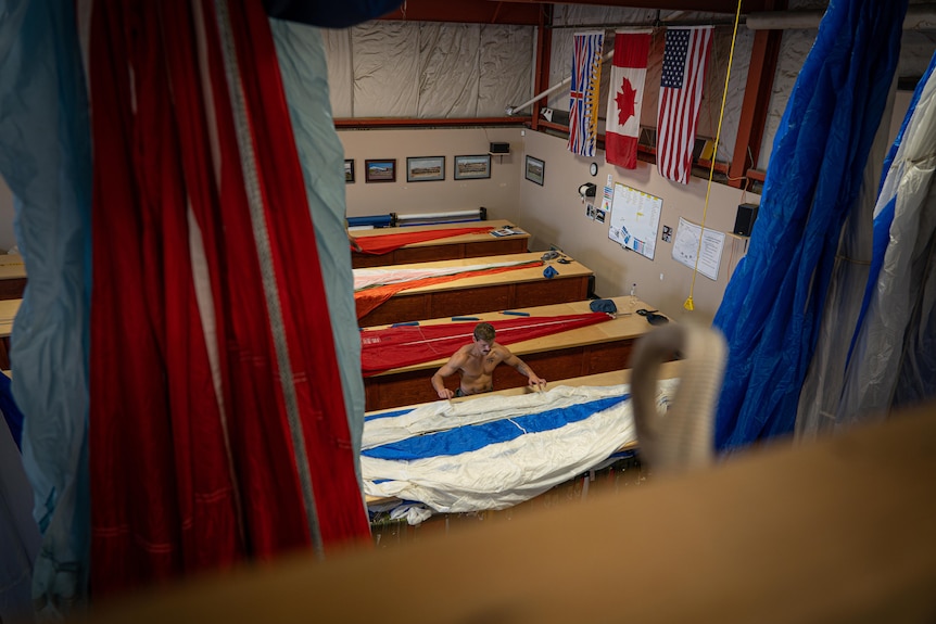 A workshop with parachutes laid out on long wooden tables. A shirtless man works on one. A Canadian flag hangs on the wall.