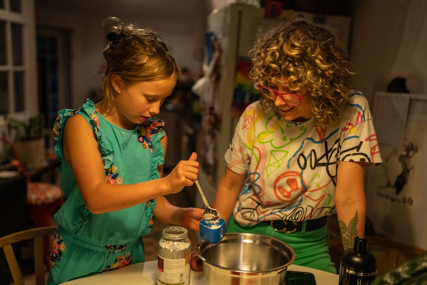 A girl holds a container and a spoon above a bowl on a kitchen counter. Her mum looks on.