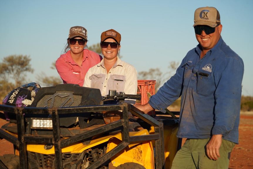 Two smiling women in caps, pink and white shirt, and a man in a cap, sunglasses, blue shirt with soil stand near a vehicle.