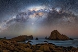 The night sky, over an ocean with rocks and twinkling stars