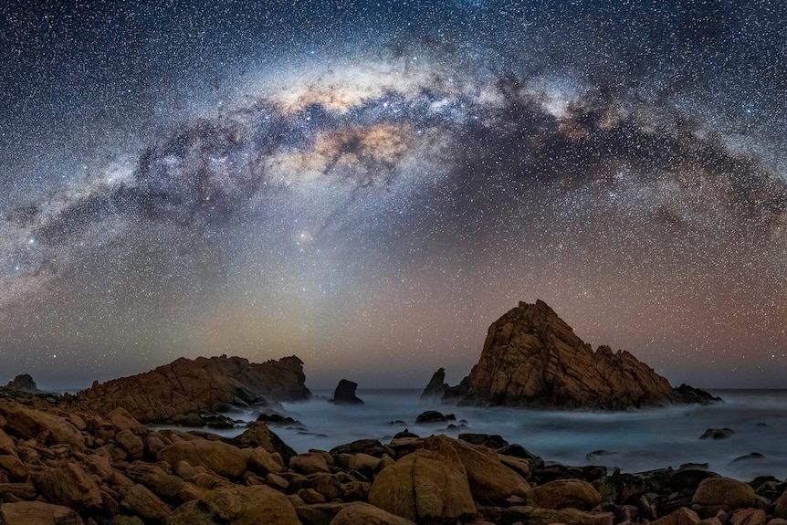 The night sky, over an ocean with rocks and twinkling stars