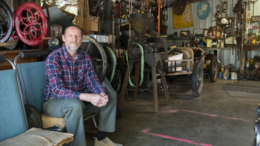 Wide shot of an older man seated in his shed with a vintage car in the background