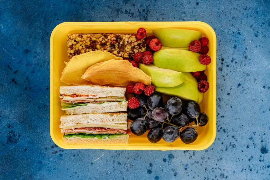 Yellow lunch box with a crust-less sandwich, fresh and dried fruit, and a muesli bar.