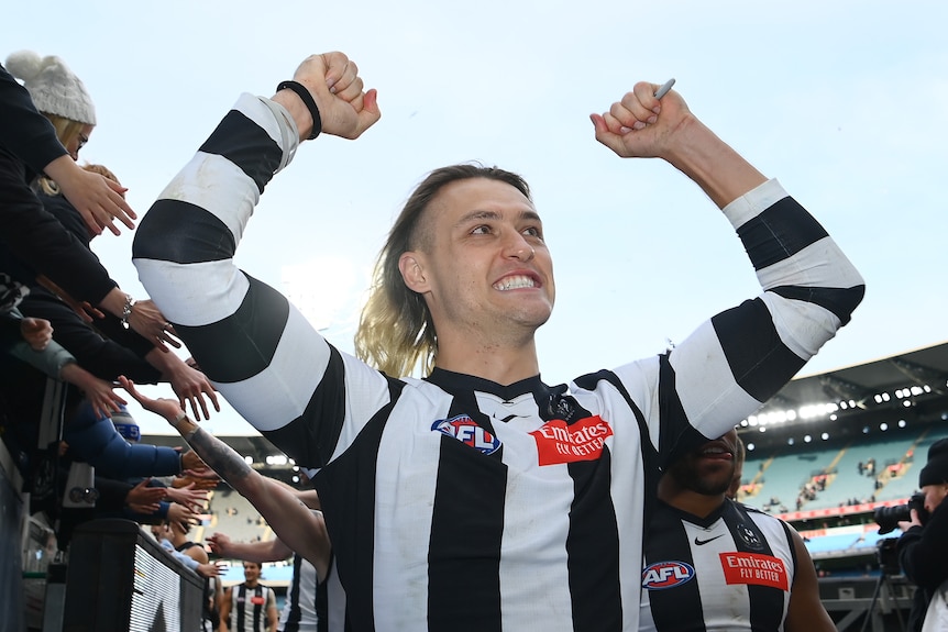 A smiling Collingwood AFL player poses with arms out as he walks down the race past fans after his team wins a game.