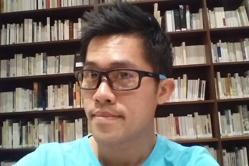 A close up of Gabriel wearing a blue tshirt and black glasses, in front of a book shelf.