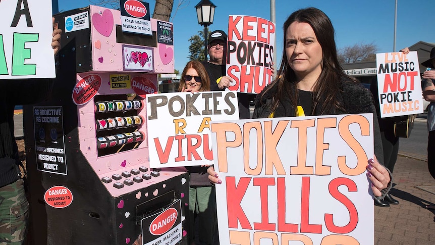 Protestors hold anti-gambling placards in front of a pink cardboard pokie machine prop