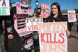 Protestors hold anti-gambling placards in front of a pink cardboard pokie machine prop