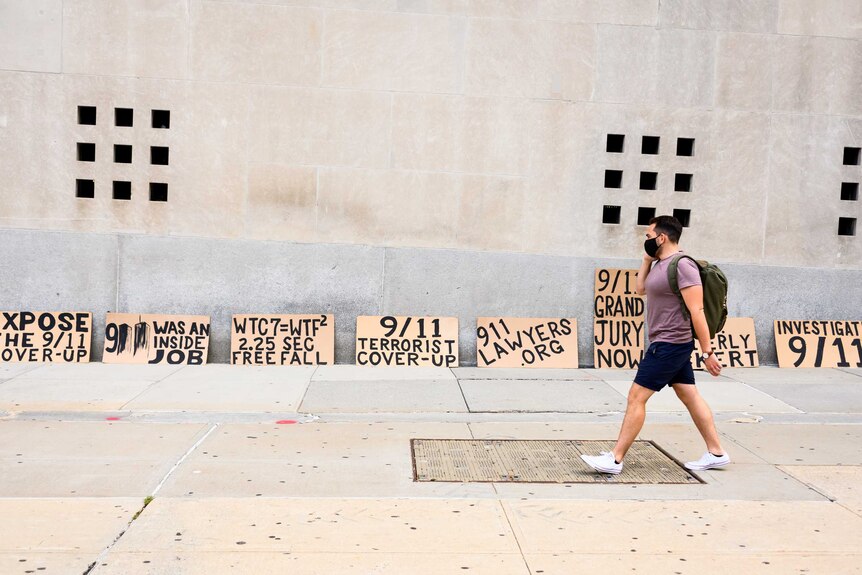 A man in a mask walks in front of a wall where signs are resting that claim 9-11 was a cover-up.