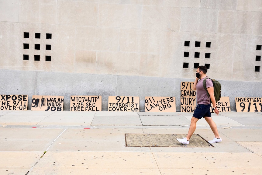 A man in a mask walks in front of a wall where signs are resting that claim 9-11 was a cover-up.