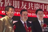 One man in a gold jacket and two men wearing suits and ties standing on stage in front of red banner