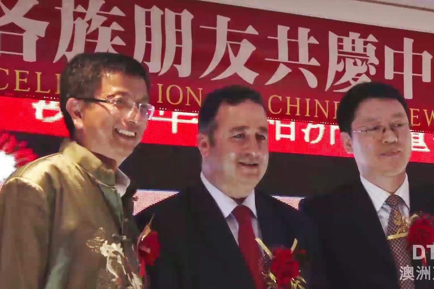 One man in a gold jacket and two men wearing suits and ties standing on stage in front of red banner