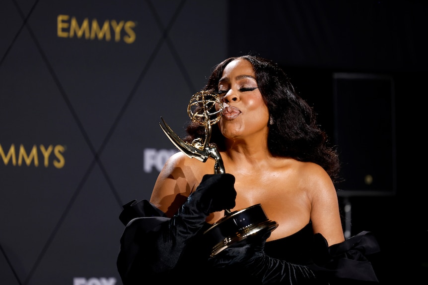 Niecy Nash-Betts holds up her Emmy Award, blowing it a kiss. She's wearing an elegant black dress.