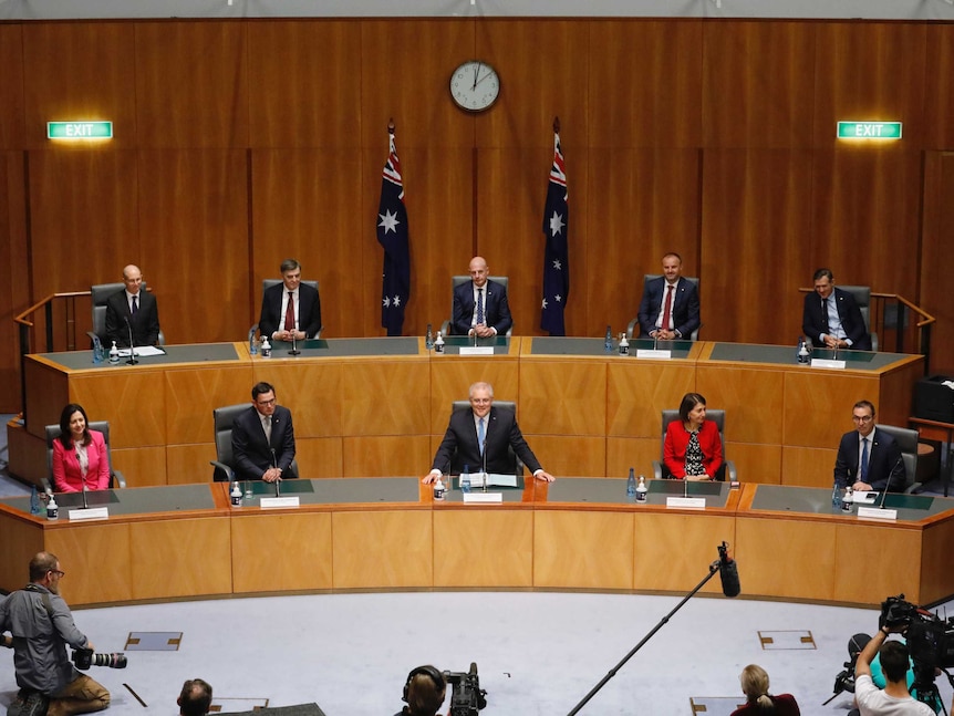 A wide shot of 10 people sitting spaced a part in a meeting hall with journalists asking them questions.