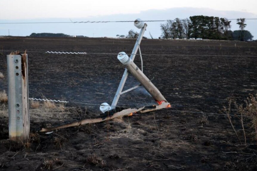 A power pole snapped in half and smouldering by a fence in a burnt out paddock.