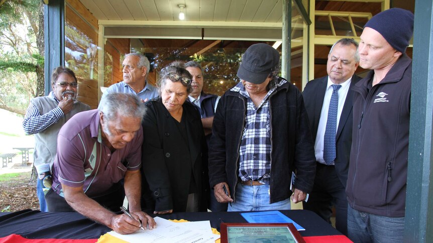 A group of people stand around a table with an Indigenous flag, signing a document