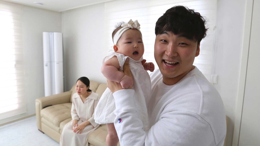A South Korean man smiles as he holds is baby in his arms while his wife sits on the couch behind him.