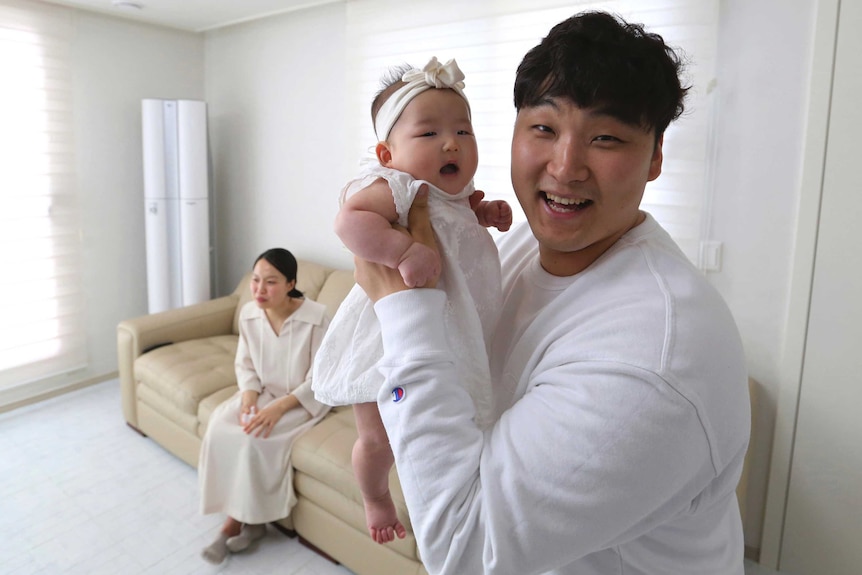 A South Korean man smiles as he holds is baby in his arms while his wife sits on the couch behind him.