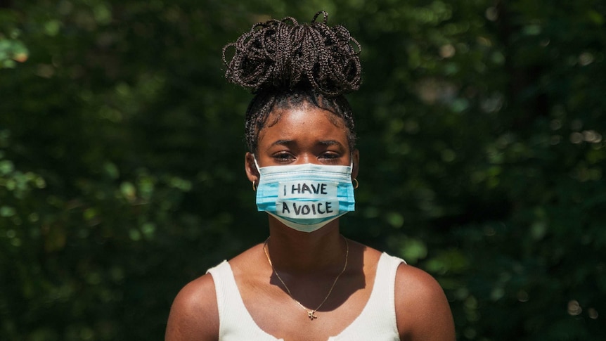 Young Black woman with plaited hair in a bun on her head. Wearing a paper mask emblazoned with the words 'I have a voice'.