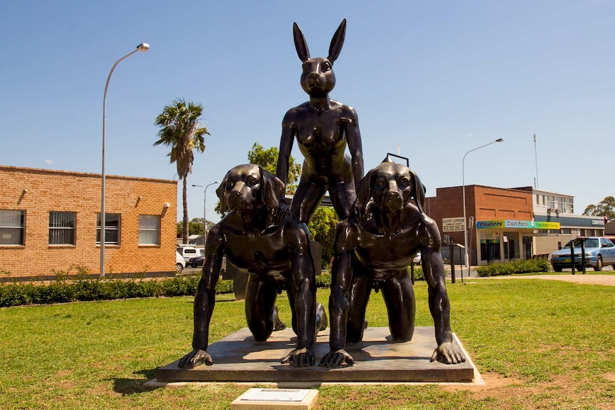 A bronze sculpture of  three human figures, one female with a rabbit head kneeling on top of two kneeling males with dog heads