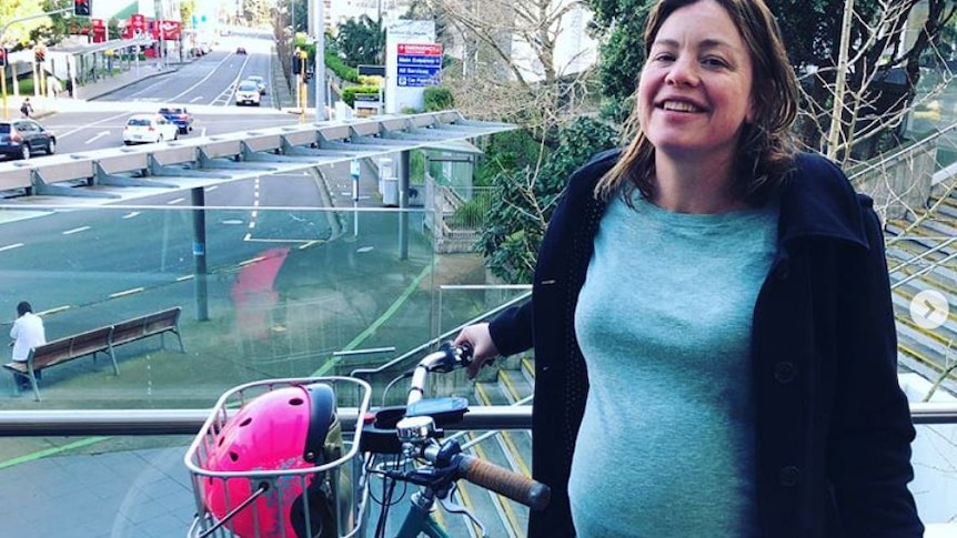 A pregnant woman stands above a main road with a pink helmet in the basket of the bicycle she holds