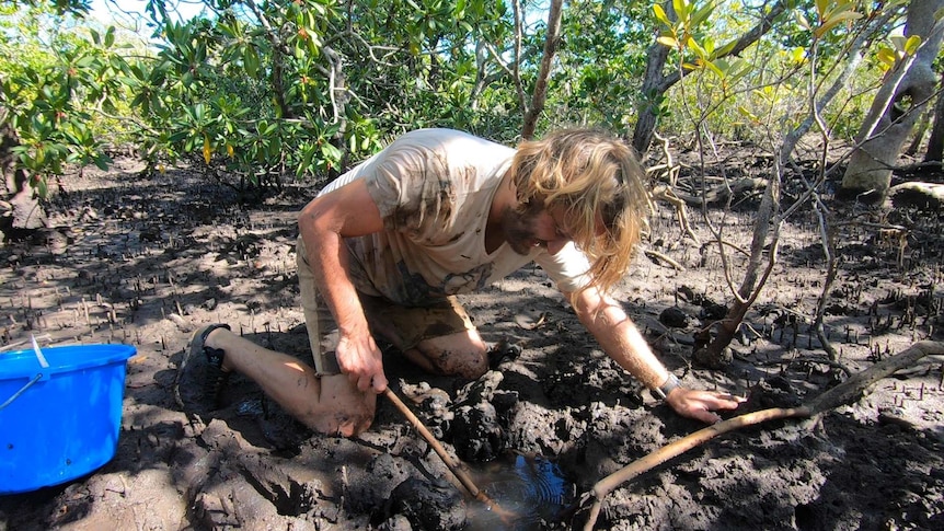 A man crouched down in the mud digging in a hole.