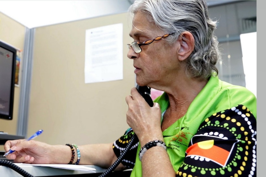 An older woman wearing a shirt decorated with Indigenous art speaks on the phone.