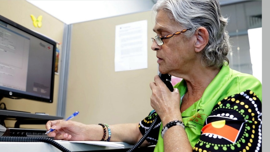 An older woman wearing a shirt decorated with Indigenous art speaks on the phone.