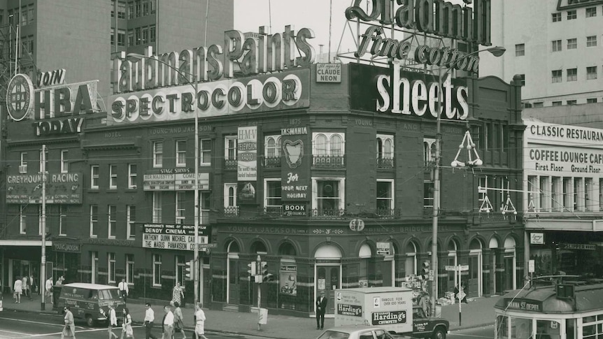 Black and white photograph of a three-story pub adorned by advertisements, on a busy city street corner.