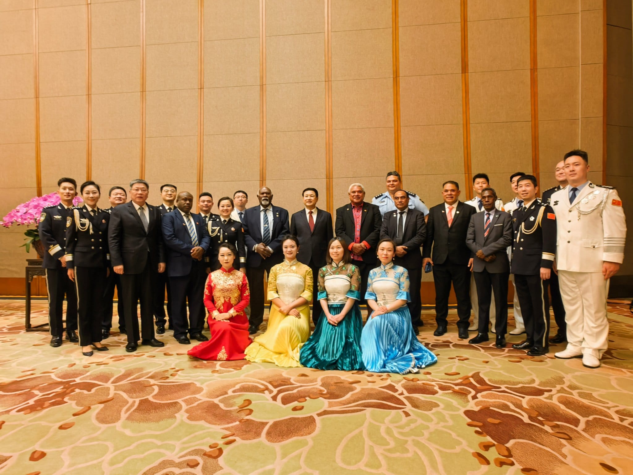 A group of women in colourful traditional Chinese ball gowns kneel in front of a row of pacific and chinese men in suits.