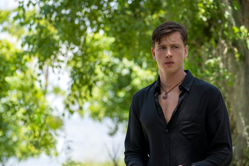 A young man in a dark button-down and a shell necklace, with a furtive expression, stands outside