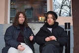 Two young women wearing winter coats and hats sit on chairs outside a coffee shop, smoking.