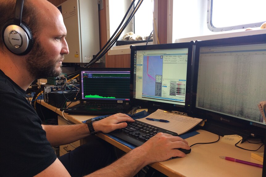A man wearing headphones sits at a computer monitoring sounds from a sonobuoy for Antarctic blue whale calls
