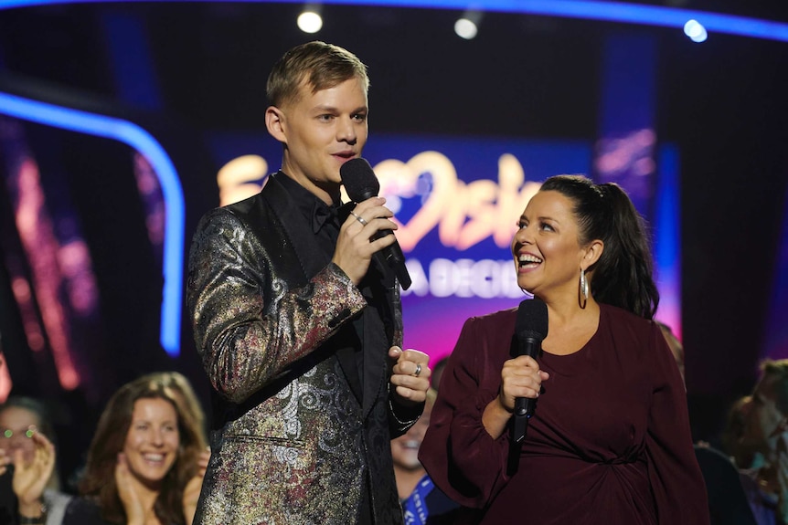 Joel Creasey stands on a stage and holds a microphone to his mouth mid-speech. Myf Warhurst is to his right. People are behind.