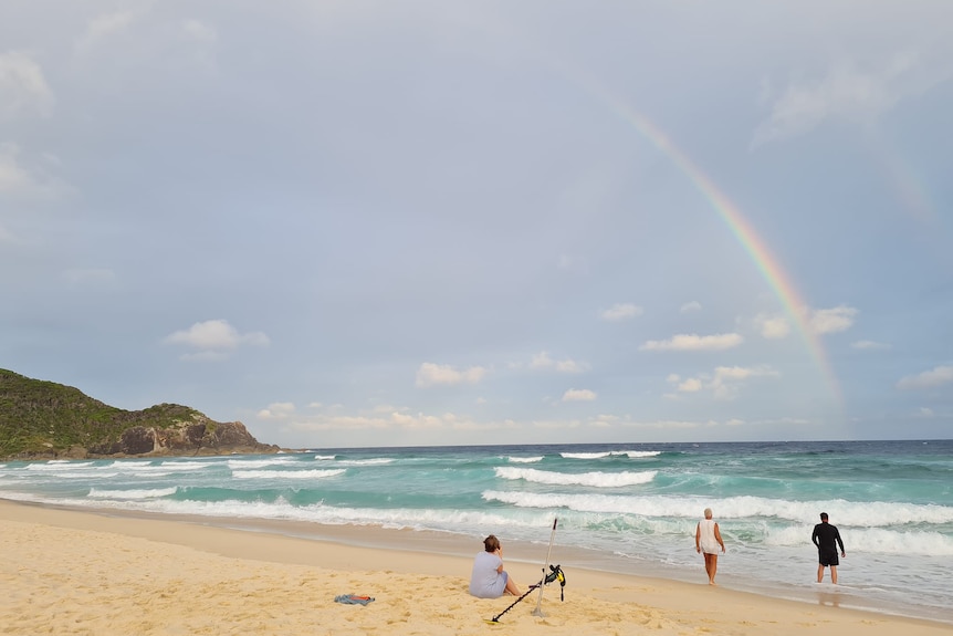 A beach, with a rainbow over the ocean and a man searching in the waves with a metal detector.