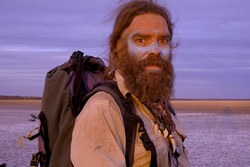 Jon Muir walks in the outback with zinc on his face, carrying a backpack.