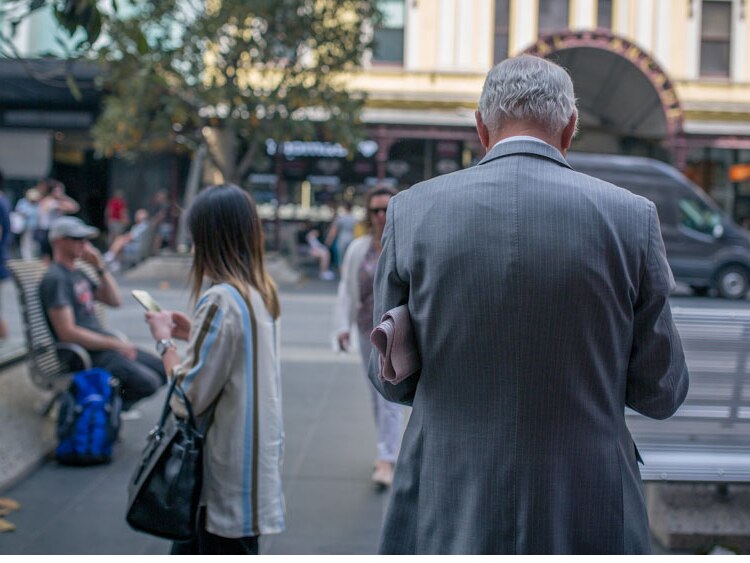 Pedestrians consume media on mobile phones as a man stands with a newspaper in Melbourne's CBD.