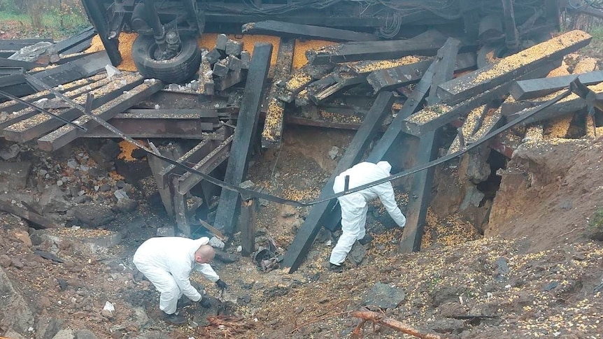 Two people in white bodysuits examine crater at site of missile explosion, with overturned truck next to them.