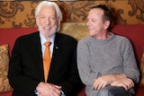 An older man with a beard wearing a suit next to another man in a grey jumper. 