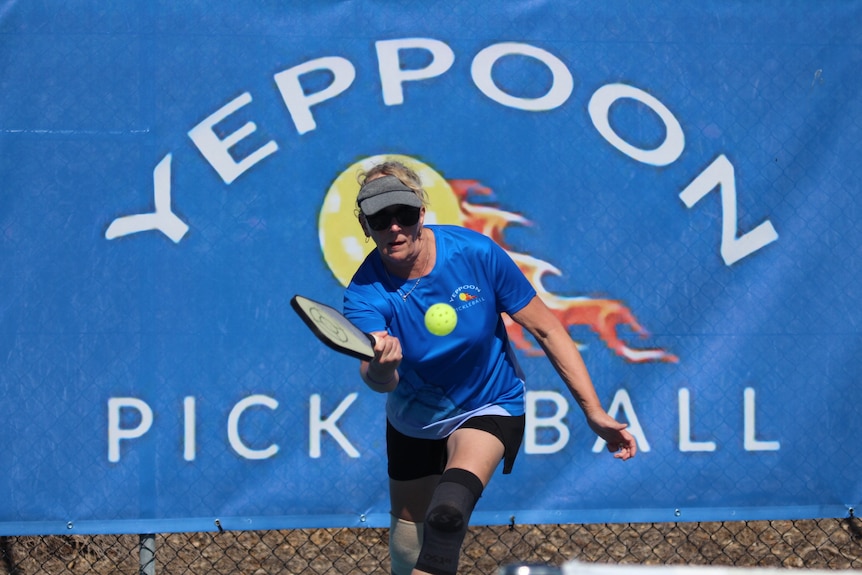A woman wearing visor, a blue t-shirt and two knee braces strikes a pickleball. 
