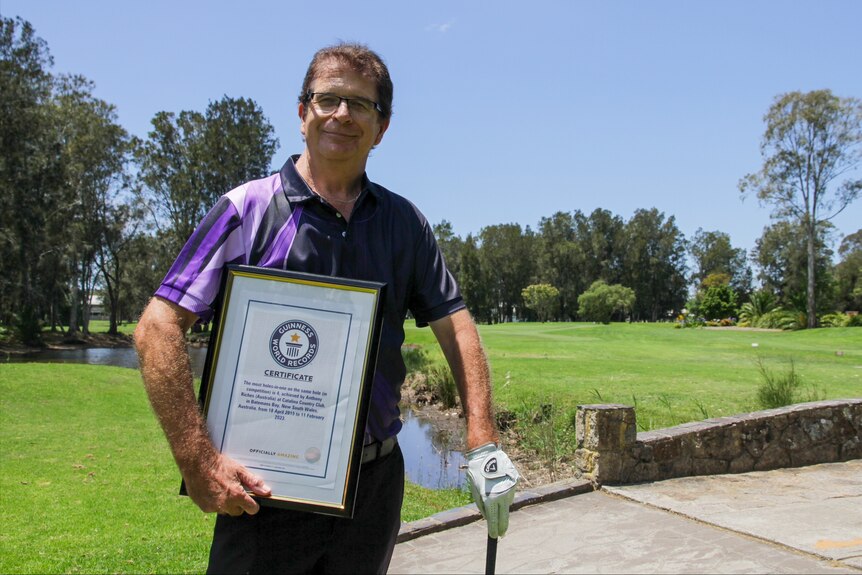 A man stands on a golf course and holds a framed certificate.