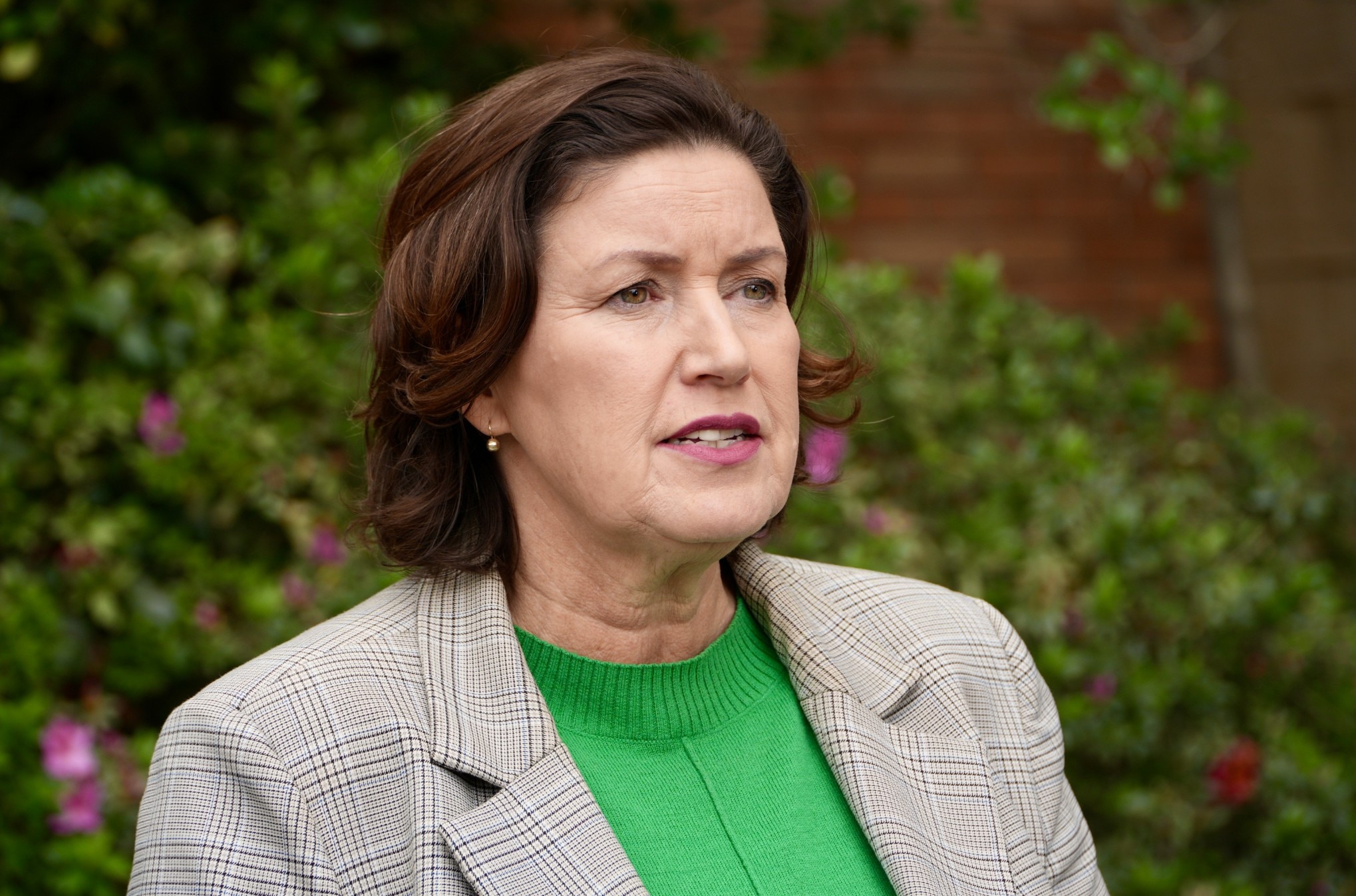 Jackie usa uma blusa verde e uma jaqueta marrom enquanto está em um palco ao ar livre.