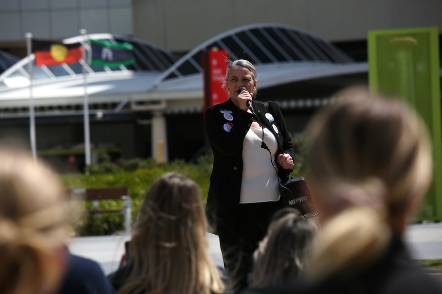 A woman wearing black and carrying a microphone speaks to a crowd of people.