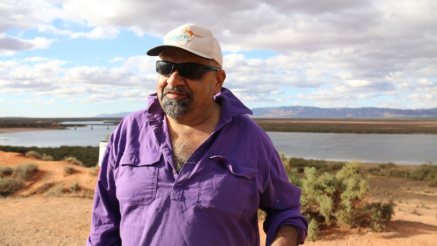 Medium portrait shot of a man in sunglasses and a purple shirt standing in front of an outback lake.