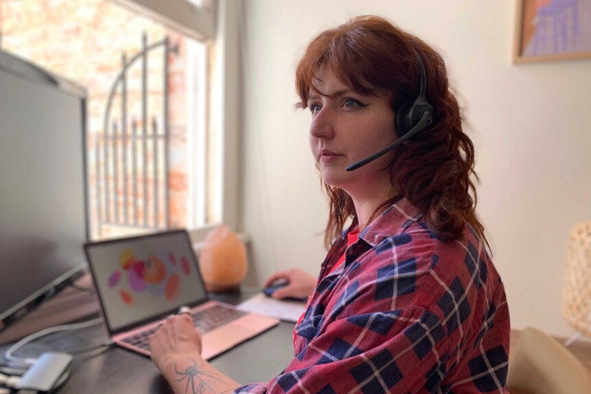 A woman with red hair, wearing a red and blue flannel shirt, sits at a computer wearing a headset.