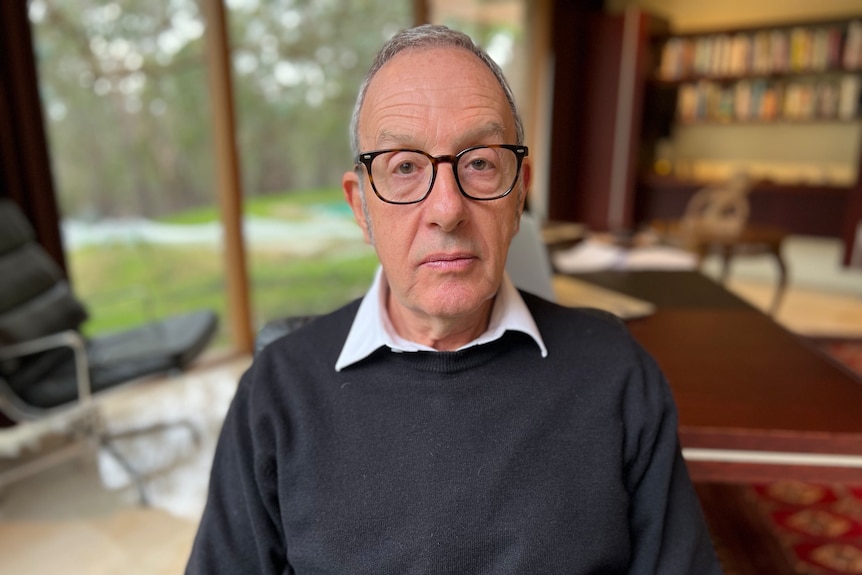 A man wearing glasses, a collared shirt and dark jumper looks into camera as he sits in an office.